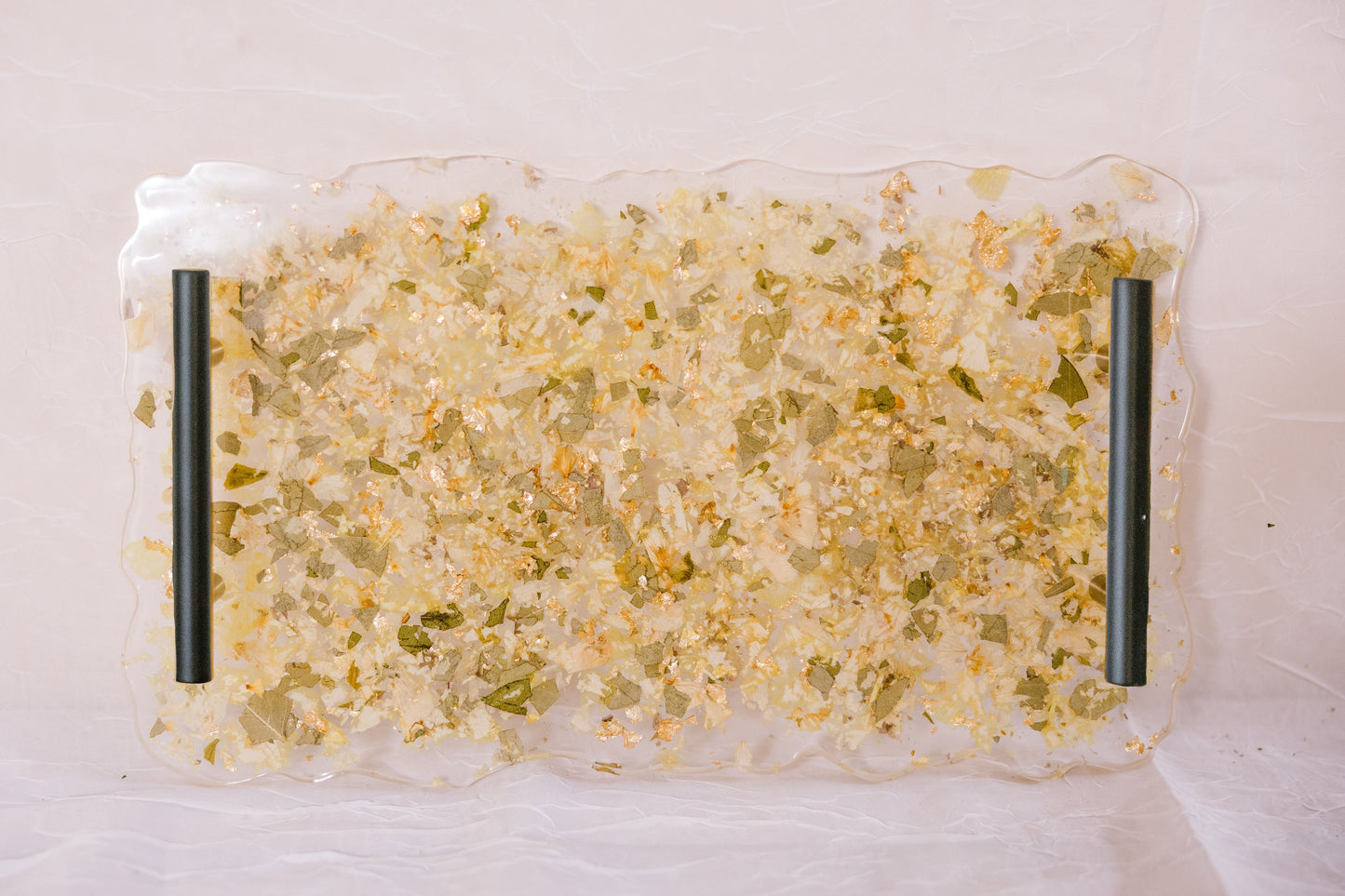 A resin serving tray with green leaves, white flowers, and gold metallic flakes designed like confetti. The tray has black handles and is against a white background. 